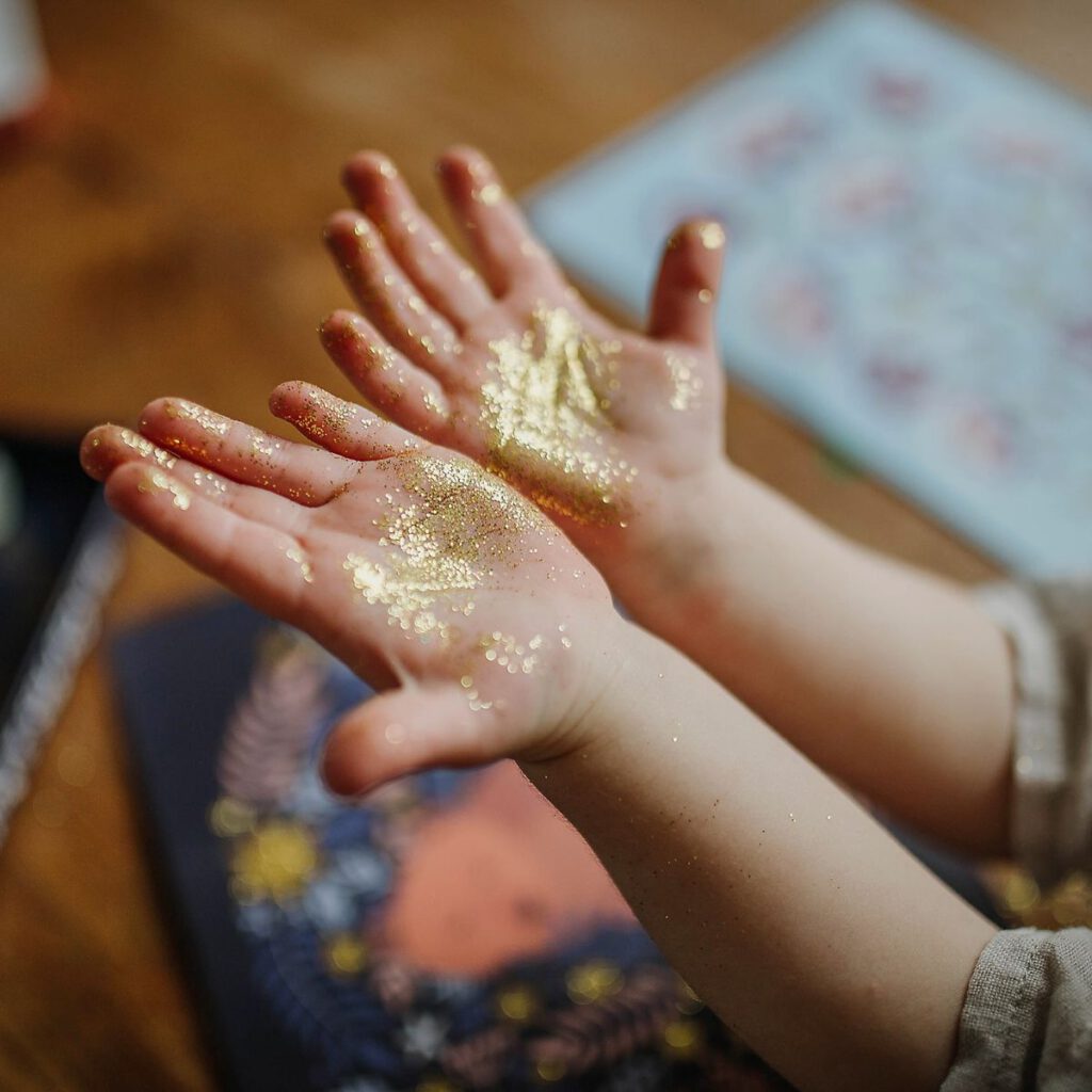 Dłonie małego dziecka ubrudzone złotym, połyskującym brokatem. W tle - tworzony właśnie projekt artystyczny DIY.