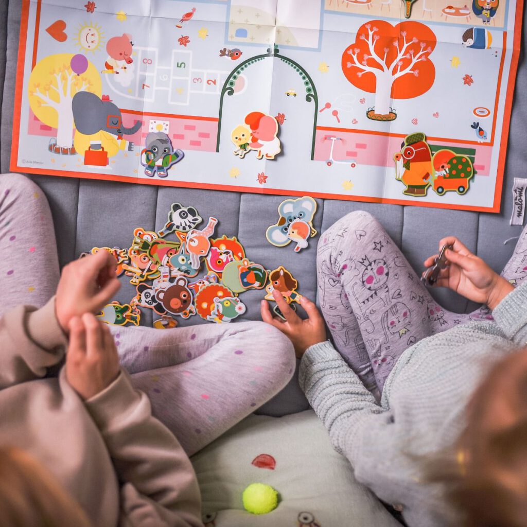 Dwie dziewczynki podczas zabawy magnesami w kształcie zwierzątek. W tle kolorowa plansza z budynkiem szkolnym.