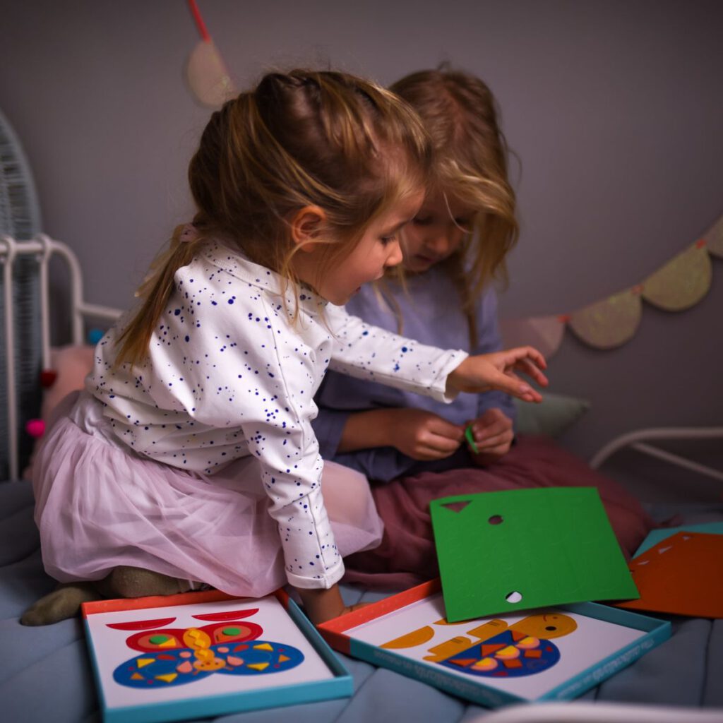 Dwie uśmiechnięte dziewczynki kreatywnie spędzają czas tworząc z piankowych, wypukłych naklejek kolorowe obrazki na planszach.