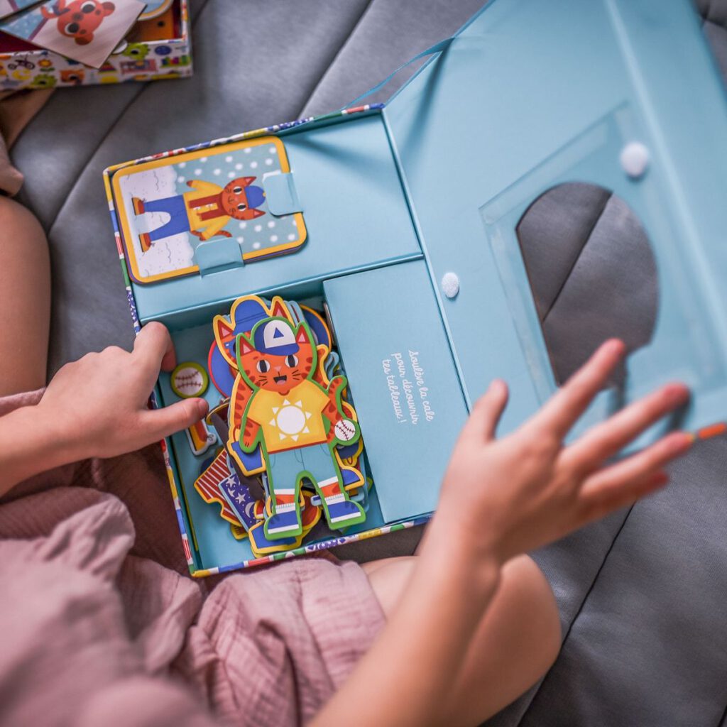 Dziewczynka otwierająca błękitne pudełko z kolorowymi magnesami w kształcie zwierzątek i elementami garderoby. 