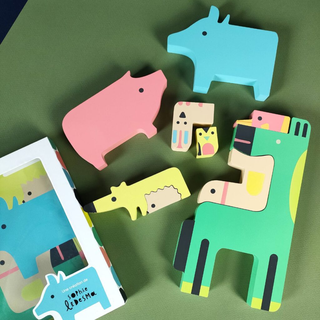 Różnokolorowe, drewniane puzzle - klocki w formie zwierzątek do układania rozłożone na oliwkowym tle. Obok pudełko.
