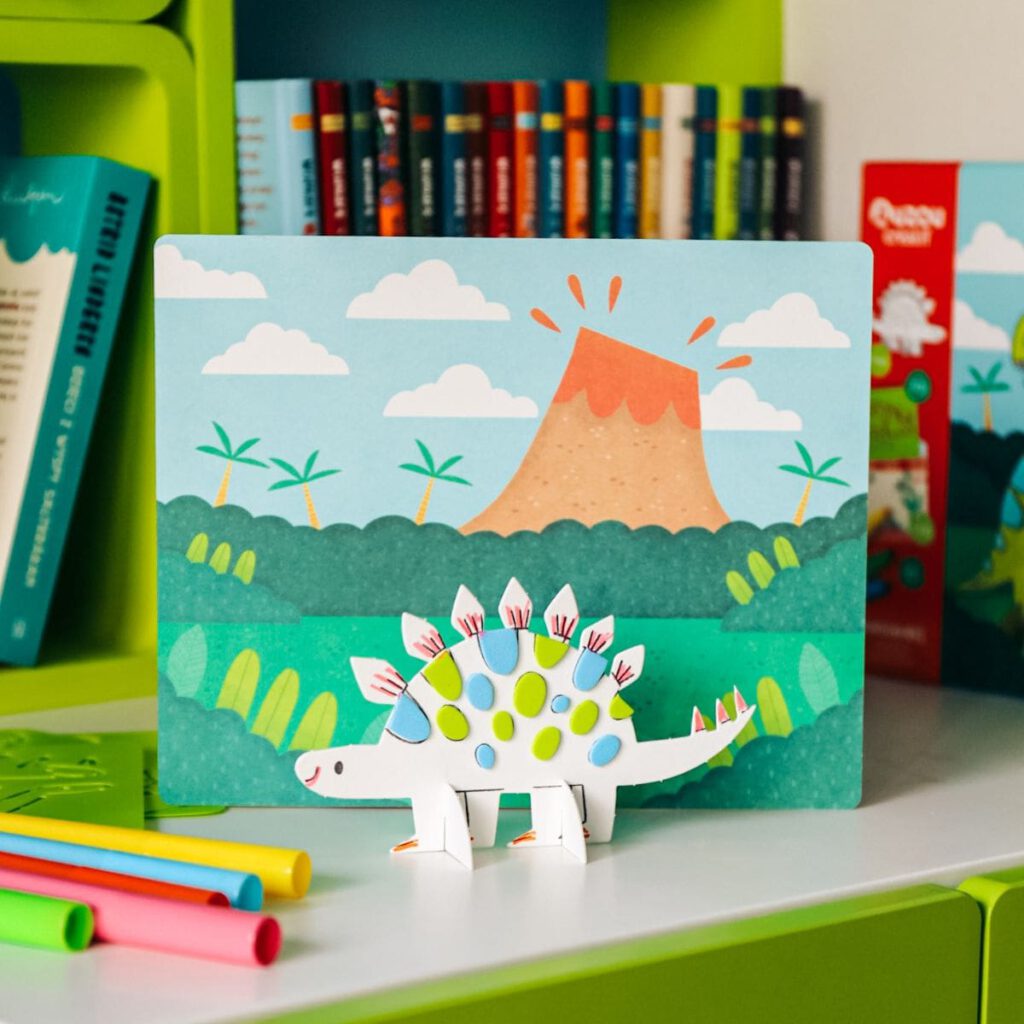 Trójwymiarowa figurka dinozaura DIY ozdobiona naklejkami, obok kolorowe flamastry. W tle plansza z zielonymi krzewami i wulkanem.
