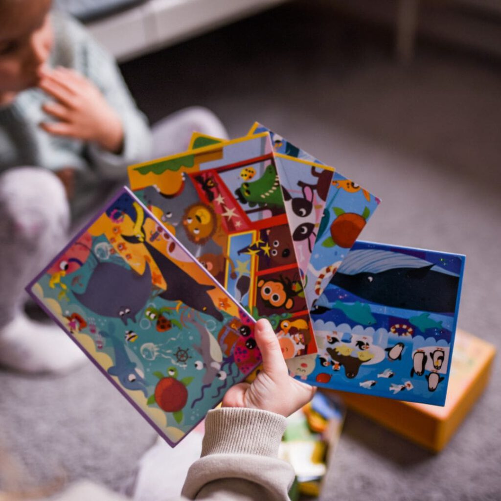 Dłoń dziecka trzymająca kolorowe plansze do gry w "Szukaj i dopasuj zwierzęta" z różnymi zwierzątkami. W tle dziewczynka i opakowanie gry.